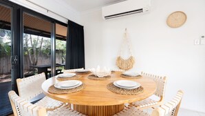 A round wooden dining table with white plates and woven placemats, surrounded by white chairs with a woven pattern.