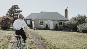 A person riding a bicycle on a path towards a stone house with a metal roof and a chimney. - Shepherd's Story, Ross (Ross)
