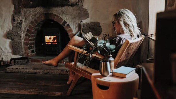 A person sitting by a stone fireplace with a book and a vase of flowers. - Shepherd's Story, Ross (Ross)