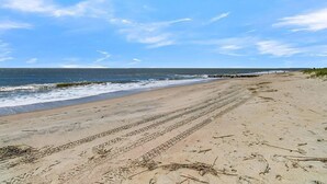 Una playa con marcas de neumáticos visibles y escombros dispersos, cielo despejado y el océano al fondo.