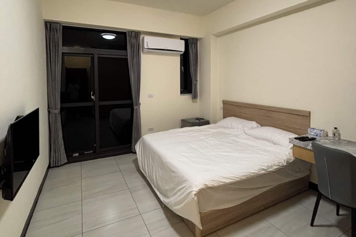 A hotel room with a bed, a desk, a chair, a TV, an air conditioning unit, and a ceiling light.