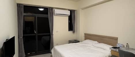 A hotel room with a bed, a desk, a chair, a TV, an air conditioning unit, and a ceiling light.