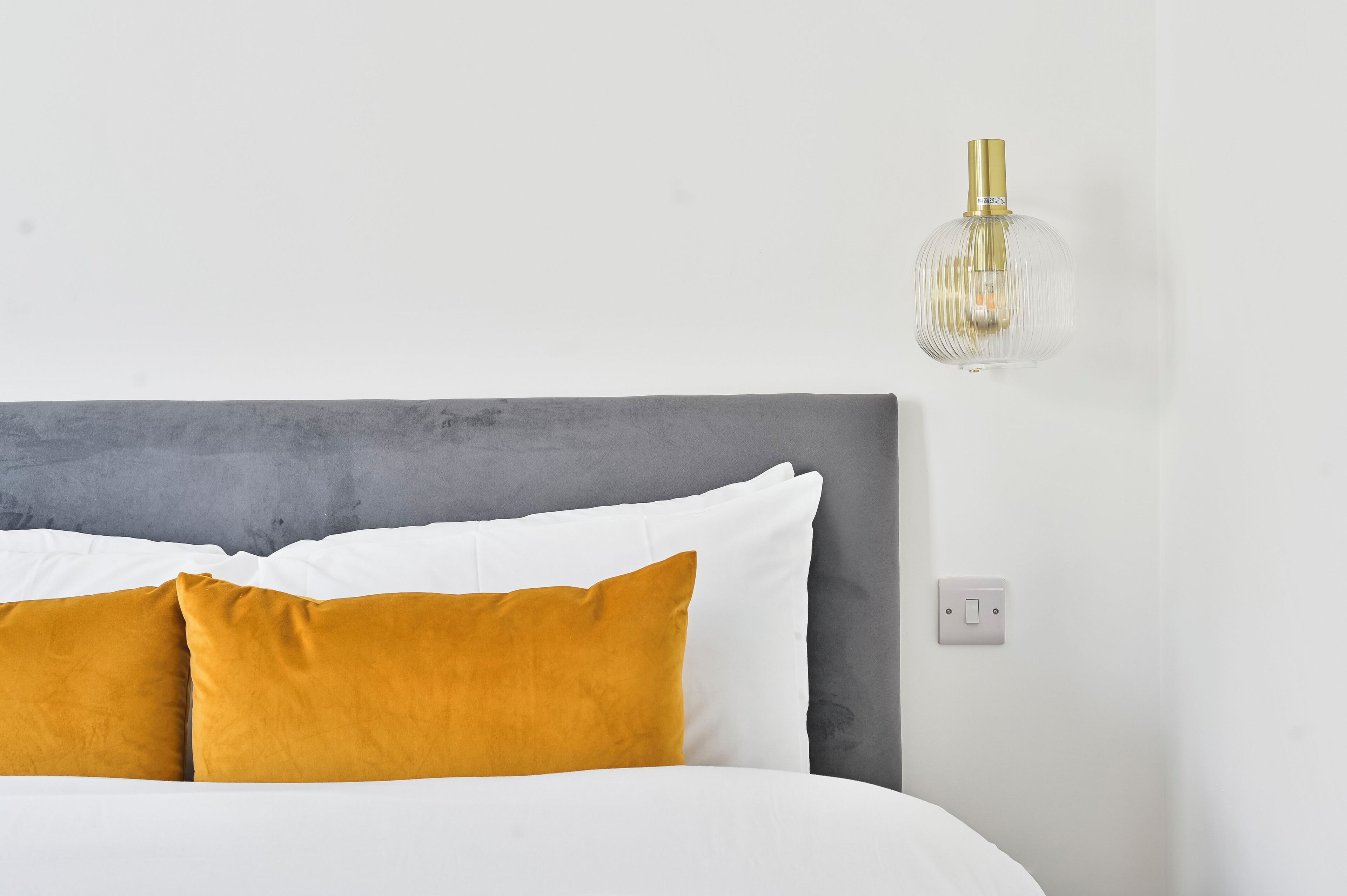 A bed with white and mustard pillows, a grey headboard, and a clear glass pendant light.