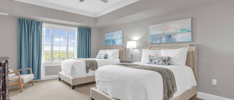 A hotel room with two beds, a ceiling fan, a large window with curtains, and artwork on the walls.