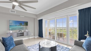 Un salon spacieux doté d’une grande fenêtre, d’un ventilateur de plafond, d’un téléviseur à écran plat et d’un balcon aménagé avec du mobilier d’extérieur.