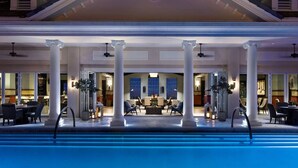Un espace aménagé au bord de la piscine, avec des colonnes, des sièges d’extérieur et une vue sur l’intérieur du bâtiment.