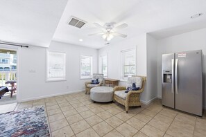 Un salon spacieux doté d’un ventilateur de plafond, d’une grande fenêtre avec stores, d’un réfrigérateur et d’un coin repas avec une table et des chaises.