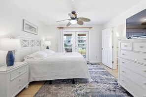 Une chambre à coucher avec un lit, des tables de chevet, une commode, un ventilateur de plafond et une vue sur l’extérieur, visible à travers une porte.