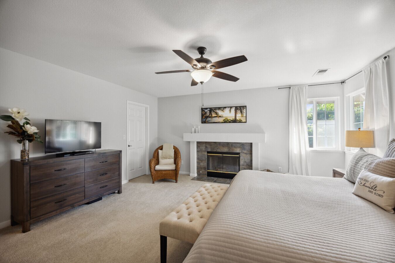 A bedroom with a bed, a TV, a dresser, a chair, a fireplace, and a ceiling fan.