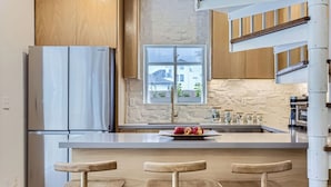 A modern kitchen with a stainless steel refrigerator, a stone backsplash, wooden cabinets, and a large window with a view of the outside.