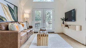 A modern living room with a brown leather sofa, a wooden coffee table, a large framed artwork, a TV, and a balcony with outdoor furniture.