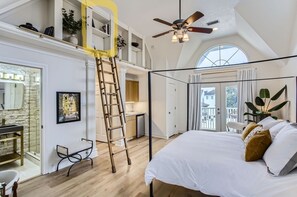 A bedroom with a large bed, a ceiling fan, a ladder, a bathroom, and a view of the outside. - Secret oasis in the heart of Montrose, outdoor dining, firepit, gazebo and grill (Houston)