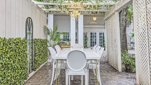 Une terrasse couverte avec une table et des chaises blanches, entourée de verdure et agrémentée d’un plafond décoratif.