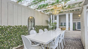 Une terrasse couverte avec une table et des chaises blanches, entourée de verdure et agrémentée d’un plafond décoratif.