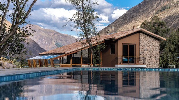 Vista de una piscina frente a una casa de fachada de piedra y tejado de tejas, con una alberca cristalina que refleja el entorno.