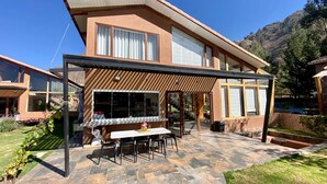 Una casa moderna de dos pisos con un patio amoblado, que incluye una mesa y sillas, rodeado de baldosas de piedra.