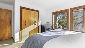 Quarto com cama, guarda-roupa de madeira, cômoda e amplas janelas com vista para árvores.
