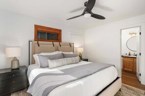 Ein Schlafzimmer mit einem Bett, Nachttischen, einem Deckenventilator, einem Fenster und einem Badezimmer mit Spiegel und Waschbecken.