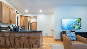 A modern kitchen with wooden cabinets, a central island with stools, a flat-screen TV, and a sofa with a cushion. - Teton Homestead: Scenic Loft Retreat (Driggs)