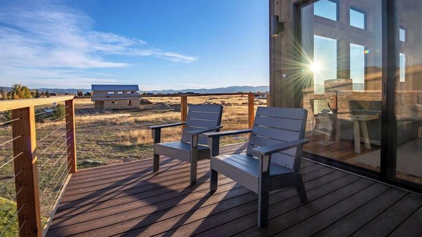 A deck with two chairs and a table, overlooking a grassy area and distant mountains. - Teton Homestead: Scenic Loft Retreat (Driggs)