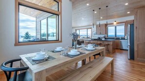 A modern kitchen with wooden cabinets, a dining area with a long table set for four, and large windows offering a view of the outdoors. - Teton Homestead: Alta Vista - Ultimate Teton Views (Driggs)