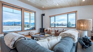 A cozy living room with a sofa, coffee table, and fireplace, large windows with a mountain view, and a lamp on a side table. - Teton Homestead: Alta Vista - Ultimate Teton Views (Driggs)