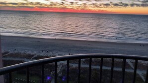 Une vue sur le coucher de soleil depuis un balcon donnant sur une plage, avec un ciel dégagé.