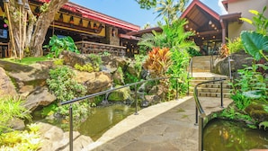 Un complexe hôtelier tropical doté d’un pont en pierre enjambant un étang, entouré d’une végétation luxuriante, et comprenant un restaurant avec un toit rouge.
