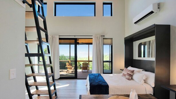 Un dormitorio con una cama, una escalera de madera, una mesa con un florero y una bandeja, y vistas al exterior.
