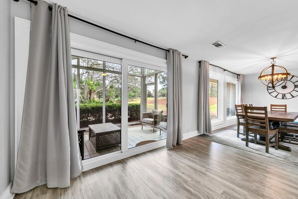 A spacious room with large sliding glass doors leading to a garden, a dining area with wooden furniture, and a modern chandelier.