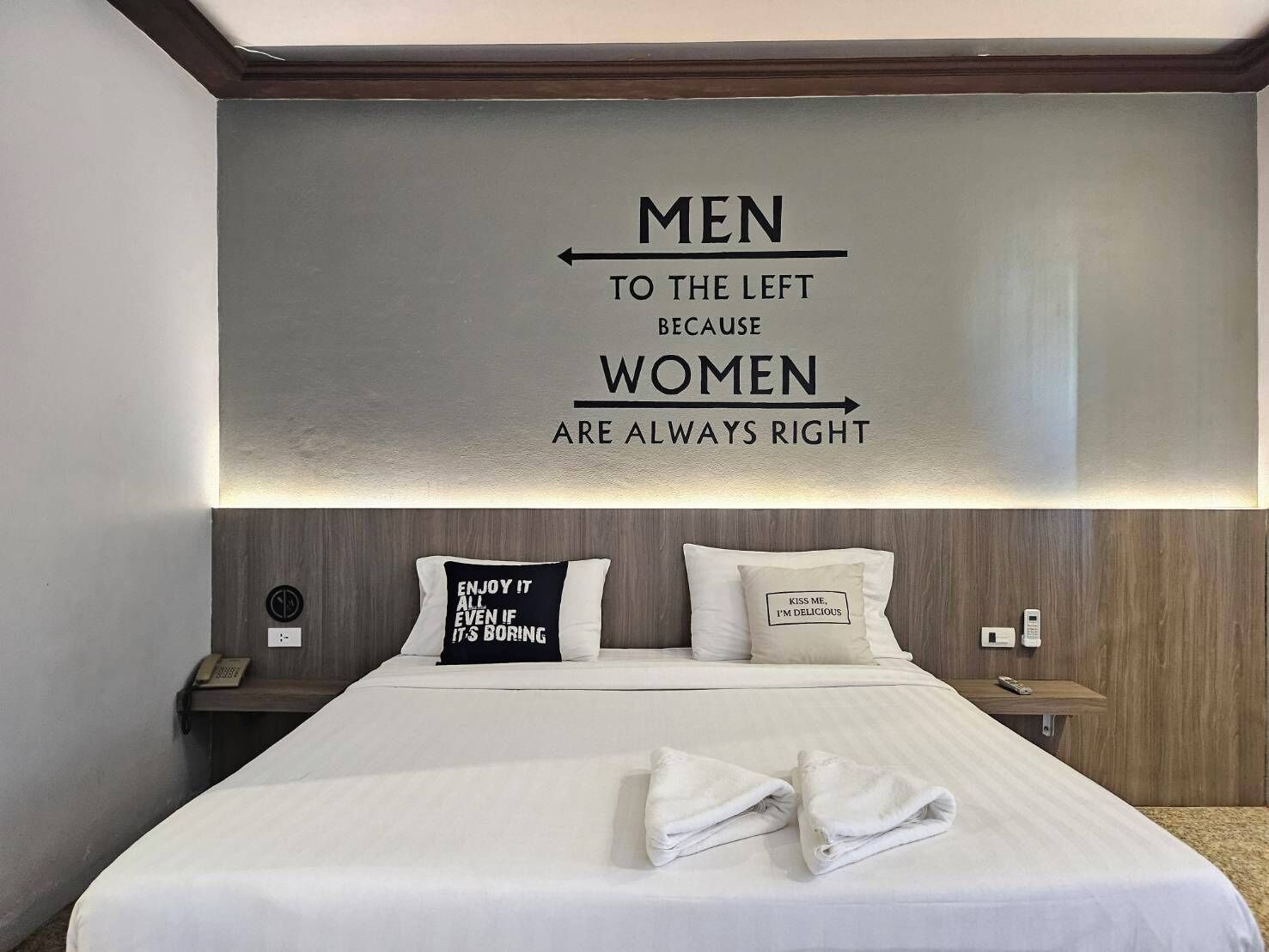 A hotel room with a bed, two pillows, a bedside table, and a wall sign.