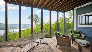 A covered outdoor deck with a hammock, wicker chairs, and a view of a lake and mountains.