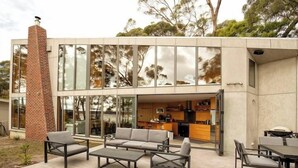 A modern house with a large glass facade, a brick chimney, and an outdoor seating area with sofas and tables. - The Beach House at Great Bay (Great Bay)