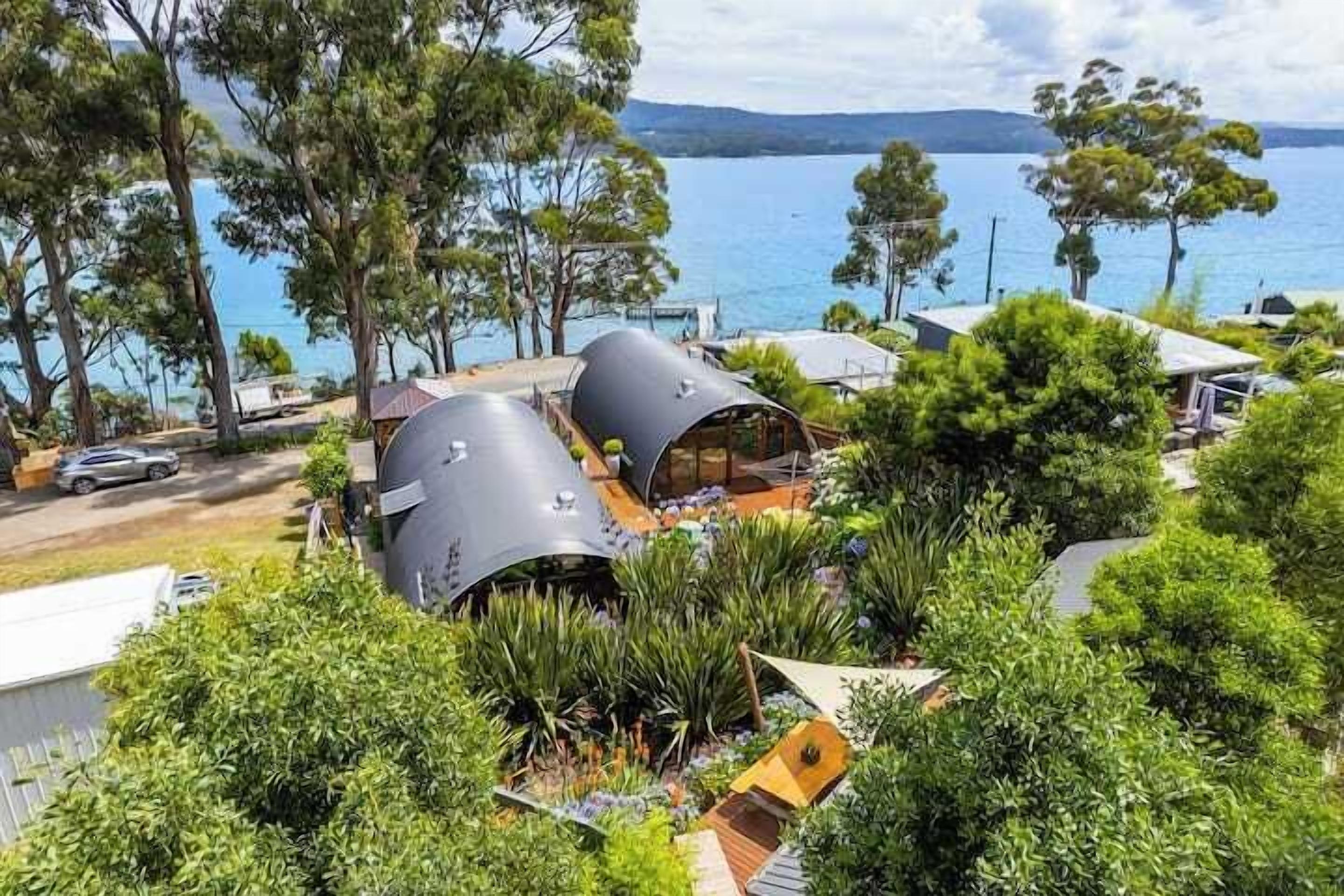 A modern eco-friendly building with cylindrical structures, surrounded by lush greenery and overlooking a body of water.