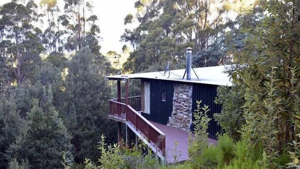 Una cabaña de dos pisos con fachada de piedra, un balcón y rodeada de un bosque denso.