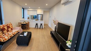 Un soggiorno moderno con un divano, un poggiapiedi nero, una TV e una cucina sullo sfondo.