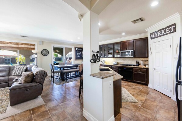 A spacious living room with a dark leather sofa and a dining area with a table and chairs. A kitchen with dark cabinets and a microwave is visible.