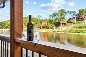 A wooden deck with a bottle and glasses, overlooking a calm body of water and a row of houses. - Near Thunder Ridge & Table Rock: Private Hot Tub, Community Pool & Free Tickets (Hollister)