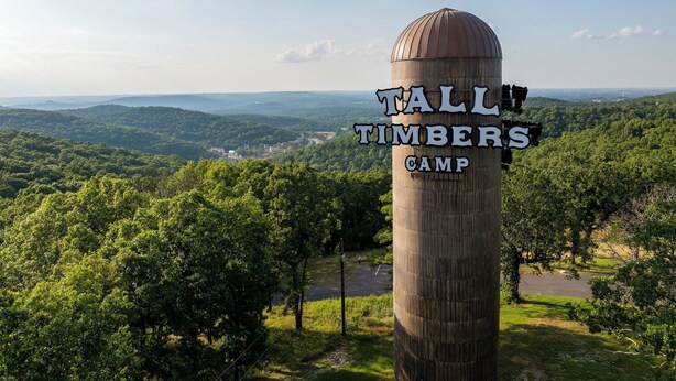 Ein hoher, zylindrischer Schild mit der Aufschrift "Tall Timbers Camp", umgeben von üppigem Grün und einer malerischen Aussicht auf weit entfernte Hügel.