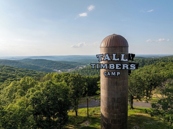 A tall, cylindrical sign with "Tall Timbers Camp" written on it, surrounded by lush greenery and a scenic view of distant hills.