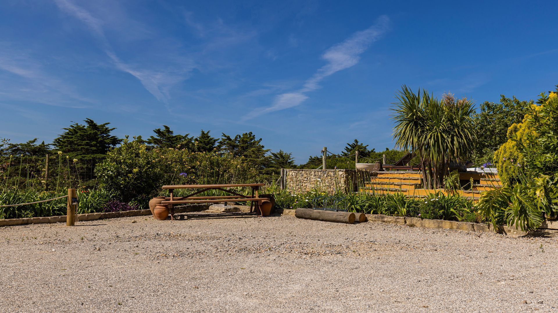 A gravel area with a wooden bench, a stone wall, and a variety of plants and trees.