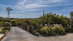 Un sendero pavimentado que conduce a una puerta, rodeado de exuberante vegetación y un molino de viento al fondo.