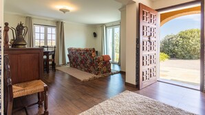 Un salón amplio con un sofá floral, muebles de madera y una puerta grande de madera que da a un jardín.
