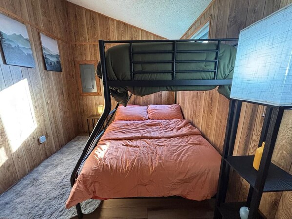 A bunk bed with a green mattress and orange bedding, wooden walls, a small window, and a wall-mounted shelf with a yellow object.