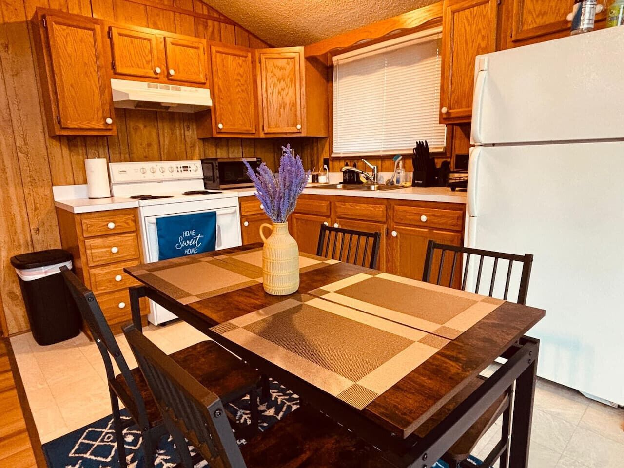Una cocina con armarios de madera, estufa, microondas, refrigerador y una mesa de comedor con sillas.