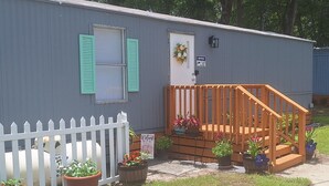 Una casa móvil con escalera de madera, cerca de piquetes blancos y plantas en macetas.