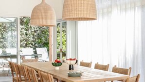 Un comedor moderno con una mesa y sillas de madera, dos lámparas colgantes de mimbre y vistas a la vegetación exterior.