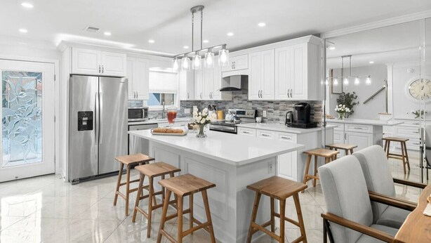 A modern kitchen with white cabinetry, a stainless steel refrigerator, a central island with stools, and a dining area with a chair and table. - Luxurious Townhome/Beach Access/11 Pools (Panama City)
