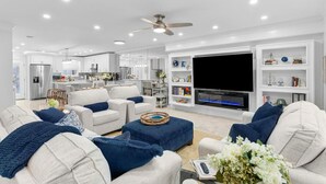 A modern living room with a large flat-screen TV, white sofas, and a kitchen in the background. - Luxurious Townhome/Beach Access/11 Pools (Panama City)