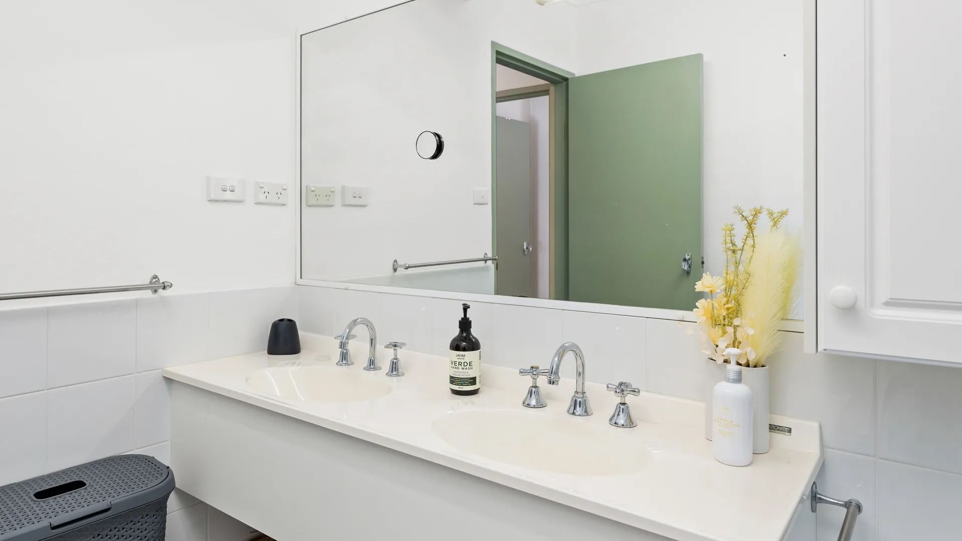 Een moderne badkamer met een witte wastafel, twee kraanwerkstukken, een spiegel en een groene deur op de achtergrond.